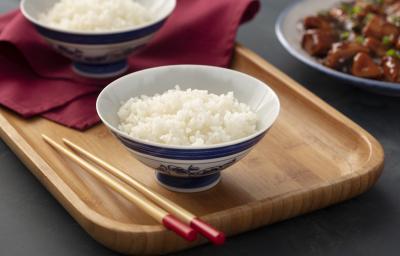 Fotografia em tons de marrom em uma bancada de madeira cinza escura com uma bandeja de madeira com um prato de cerâmica branca com detalhes em azul com o arroz japonês dentro dele. Ao lado, hashi e um pano na cor vinho.