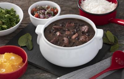 Fotografia em tons de vermelho em uma bancada de madeira escura, uma tábua preta com uma panela branca com a feijoada dentro dela. Ao lado, vários potinhos com laranja, couve, arroz e farofa.