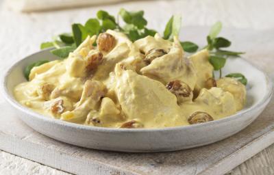 Fotografia em tons claros de uma salada de frango com molho ao curry e algumas folhas em um recipiente de cerâmica raso na cor branco. O prato está sobre uma peça de madeira em tom claro.