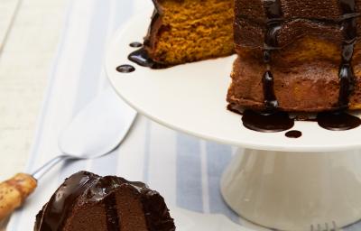 Fotografia em tons de marrom em uma bancada de madeira com um pano branco com listras azuis. Ao centro, um prato branco redondo raso com uma fatia do bolo de cenoura com chocolate. Ao fundo, um suporte branco para bolos com o bolo inteiro.