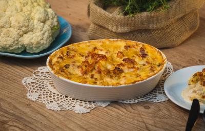 Fotografia em tons de amarelo em uma bancada de madeira clara, um paninho de renda e um recipiente branco oval com a couve-flor gratinada feita na AirFryer Mondial dentro dele. Ao lado, um prato com couve-flor crua, um outro prato com uma fatia.