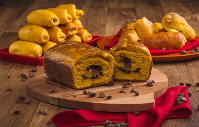 Foto de uma bancada de madeira. Sobre ela há uma tábua sobre um pano vermelho e nela está um pão em tom laranja aberto ao meio com recheio de chocolate. Ao fundo há mais pães menores e espalhadas pela bancada estão algumas gotas de chocolate