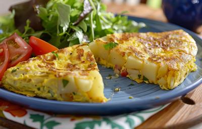 Foto aproximada da receita de omelete com batata, servida em um prato e dividia ao meio, podendo ver as batatas no centro. No prato há uma salada verde e algumas fatias de tomate