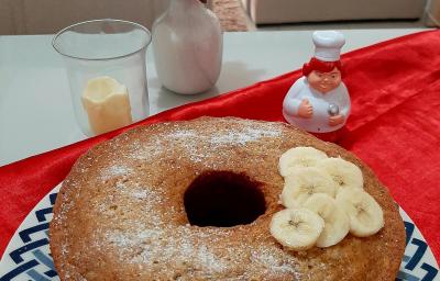 Fotografia em tons de azul em uma mesa com um pano vermelho, um prato redondo raso com desenho em azul e um bolo de banana com canela e rodelas de banana em cima dele.