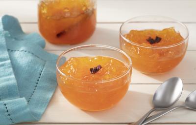 Fotografia em tons de laranja em uma bancada de madeira branca, um pano azul, três potinhos de vidro com o doce de mamão ralado e em pedaços pequenos com cravos. Ao lado, duas colheres.