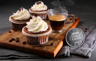 Fotografia em tons de marrom em uma bancada cinza com pano cinza escuro, uma tábua de madeira com três forminhas com cupcakes feitos com café e leite dentro delas. Ao lado, uma xícara de café e a cápsula do Café Barista Dolce Gusto.