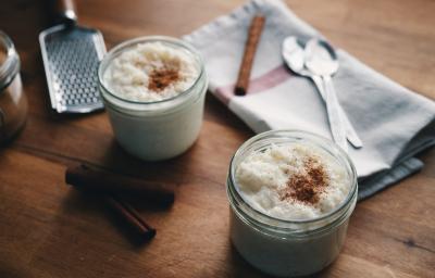 Fotografia de dois potes pequenos de vidro com arroz doce e canela polvilhada por cima. No fundo da foto, duas colheres de sopa e uma canela em pau sobre um pano de pano branco com uma listra vermelha. Ao lado dos potes, tem duas canelas em pau.