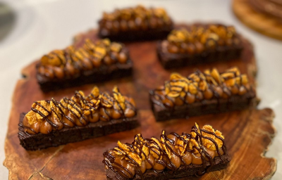 Fotografia em tons de marrom com uma bancada de madeira ao centro. Em cima da bancada existe 6 sobremesas com base de brownie e cobertas com um creme de caramelo e chocolate