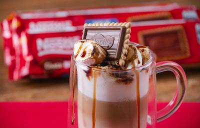 Fotografia em tons de vermelho com uma caneca de vidro ao centro. Dentro da caneca existe uma receita de café com leite finalizada com marshmallow e Choco Biscuit NESTLÉ