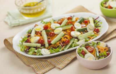 Fotografia em tons de branco, rosa e verde, com saladeira ao centro contendo folhas verdes, vegetais e ovo de codorna sobre guardanapo colorido, no entorno cumbucas rosa e verde com salada, ao fundo potinho com azeite, tudo sobre bancada na cor rosa.