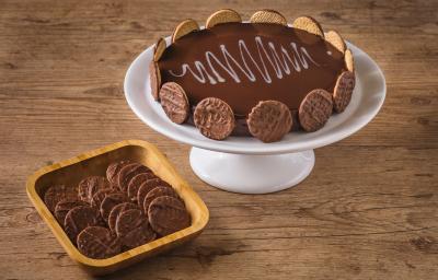 Imagem de uma torta decorada com biscoitos ao redor, disposta num prato branco alto, ao lado de uma tigela de madeira com mais biscoitos dentro