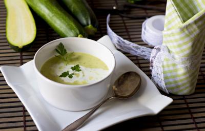 Fotografia de um recipiente fundo branco de vidro com uma sopa de abobrinha e cará, com um creme branco por cima e salsinha. A sopa está em cima de um prato de vidro branco e quadrado, com uma colher de sopa apoiada nele, sobre uma mesa de madeira.