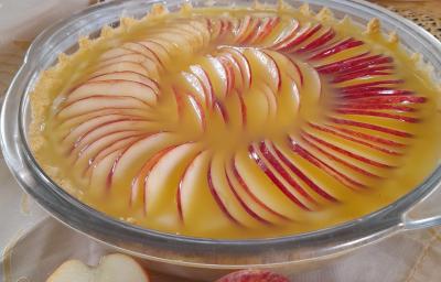 Foto da receita de Torta de Maça Dona Kilza. Observa-se um recipiente refratário transparente redondo e grande com a torta. A cobertura é feita de maçãs laminadas finas dispostas em círculos com um creme brilhoso de laranja.