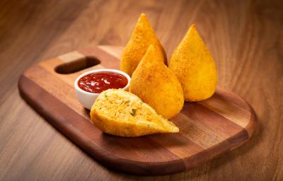 Fotografia em tom amadeirado com uma tábua de madeira ao centro. Em cima da tábua existe 3 coxinhas douradas e outra cortada pela metade recheada com frango desfiado. Ao lado existe um pequeno pote cheio de ketchup.