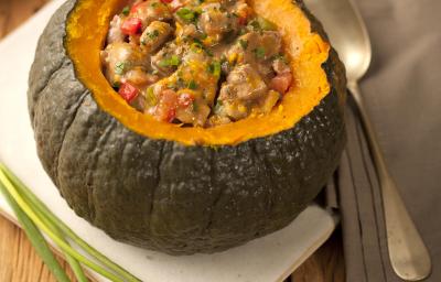 Fotografia em tons de marrom e cinza de uma bancada de madeira com um paninho branco, sobre ele uma abóbora recheada com carne e legumes. Ao lado cebolinhas e um paninho cinza com uma colher, ao fundo um prato branco.