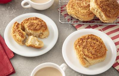 Foto da Receita de Coconut Rolls. Observa-se dois pratos de sobremesa com duas unidades de coconut rolls cada. Atrás, tem uma grelha com mais duas unidades. Duas xícaras de café com leite e toalhas vermelhas decoram a foto.