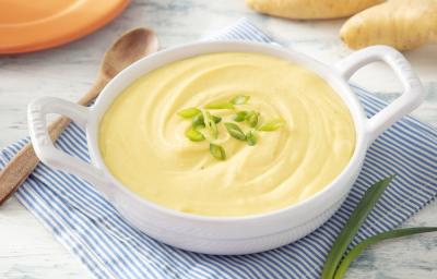 Foto em tons de amarelo da receita de purê de mandioquinha zero lactose servida em uma panela branca sobre um pano listrado azul e branco com uma colher de madeira ao lado