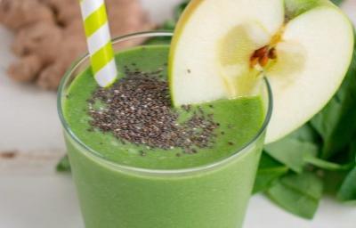 Fotografia em tons de verde em uma mesa branca com um copo de vidro alto e o smoothie de maçã verde com banana e chia dentro dele, com uma fatia de maçã enfeitando o copo. Ao fundo, folhas de espinafre e gengibre.