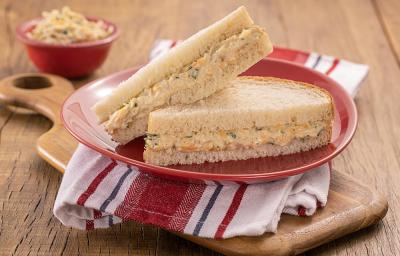 Foto da receita de sanduíche com patê de frango servido em duas porções juntas em um prato vermelho sobre um pano branco e vermelho listrado. Embaixo uma tábua de madeira e ao fundo um recipiente vermelho com o frango desfiado para decoração