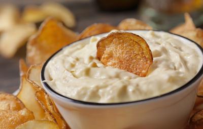 Fotografia de um recipiente pequeno de vidro com um patê de creme de leite, ketchup e creme de cebola com um chip de batata por cima. Em volta e no fundo, mais chips de batata sobre um apoio preto.