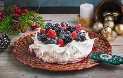 Foto da receita de pavlova natalina, branca, sobre um prato vermelho, decorada com frutas vermelhas e açúcar. Ela está numa mesa de madeira preenchida de decorações de natal como pinhas, bolas de natal e uma vela acesa ao fundo