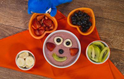 Fotografia em tons de laranja em uma bancada de madeira escura, um pano laranja, um recipiente redondo com o smoothie de morango, banana e kiwi dentro dele e frutinhas formando um rostinho feliz. Ao lado, potinhos com as frutas e o ursinho azul de pelúcia