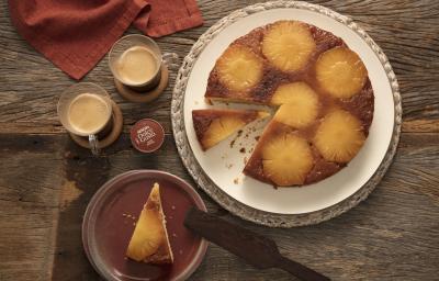 Foto de cima de um bolo invertido com rodelas de abacaxi, uma fatia está servida em um prato de sobremesa ao lado e duas xícaras transparentes com café preto e uma cápsula de Dolce Gusto Café Matinal.