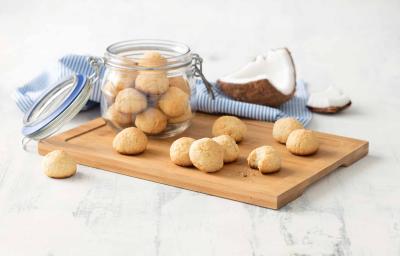 Receita em tons de bege e azul de uma bancada branca vista de cima. Contém uma tábua bege com um pote transparente com biscoitinhos dentro e por cima da tábua contém mais biscoitos. Ao fundo um pano azul e uma coco cortado.