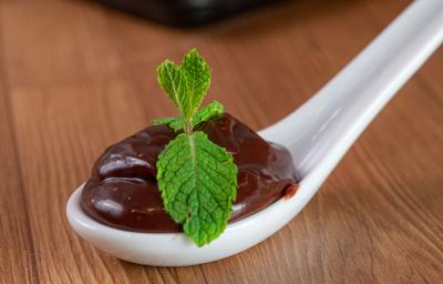 Fotografia de uma colher de servir branca de vidro, com uma porção de brigadeiro com duas folhas de hortelã por cima. A colher está apoiada sobre uma mesa de madeira.