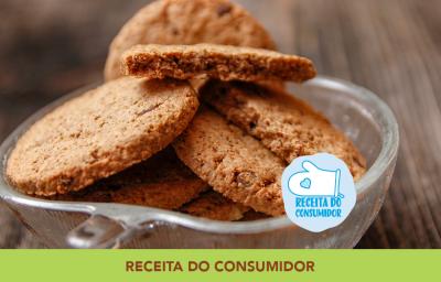 Foto aproximada da receita de Cookies Integrais, dentro de um pote de vidro, em tom marrom claro, sobre uma bancada de madeira e com dizeres no rodapé em tom verde.