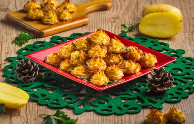 Foto da receita Batatas Natalinas, em tom amarelo e dourado, servidas em um prato vermelho quadrado sobre uma toalha verde natalina. Tudo está em uma mesa de madeira decorada com batatas cruas, pinhas natalinas e mais algumas batatas prontas.