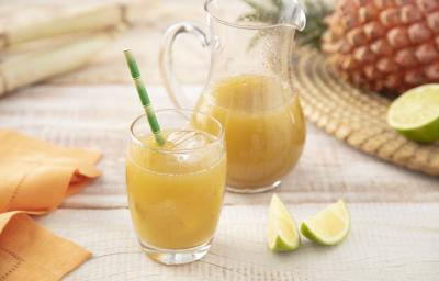 Fotografia em tons de laranja e verde em uma bancada de madeira clara, um pano laranja, um jogo americano de palha com um abacaxi e limão em cima dele. Ao centro, um copo de vidro com caldo de cana dentro e uma jarra com o restante do caldo de cana.