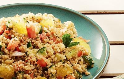 Fotografia em tons de verde e amarelo, com porcelana verde contendo tabule de quinoa com legumes e gomos de laranja, decorado com folhas de hortelã, tudo sobre bancada de madeira bege.