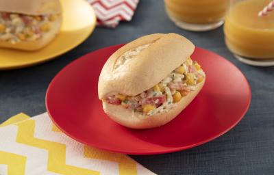 Foto da receita de sanduíche saudável sem glúten, montado em um pão francês recheado com um creme de frango, milho, sobre um prato vermelho e uma mesa com tecidos em tons amarelo e vermelho, ao lado de dois copos de suco e mais um sanduíche em outro prat