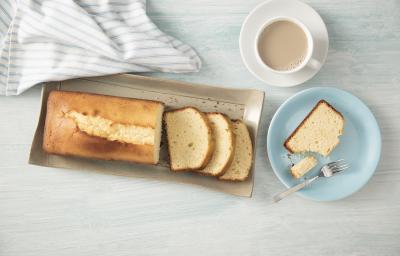 Fotografia em tons de azul em uma bancada de madeira clara com um prato redondo raso azul com uma fatia de bolo de Leite Moça. Ao lado, um recipiente retangular bege com o bolo inteiro.