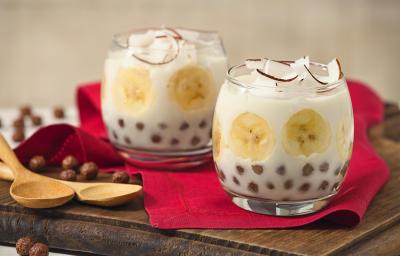 Fotografia em tons de vermelho e marrom em uma bancada branca com uma tábua de madeira escura, um pano vermelho, duas taças de vidro com o docinho de banana com Nescau Cereal.