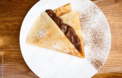 Fotografia vista de cima de uma panqueca com calda quente de mel e chocolate. A panqueca está em um prato branco raso com um pouco de cacau em pó peneirado por cima. O prato está sobre uma mesa de madeira.