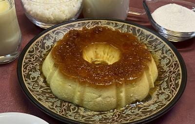 Foto em tons de bege escuro da receita de pudim de arroz inteira servida sobre um prato grande decorado. Ao fundo há alguns recipientes com arroz, açúcar, leite ninho e leite MOÇA.