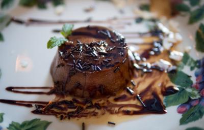 Fotografia de um mini pudim de chocolate sem furo no meio, decorado com granulado, calda de chocolate e uma folha de hortelã, sobre um prato branco desenhado..