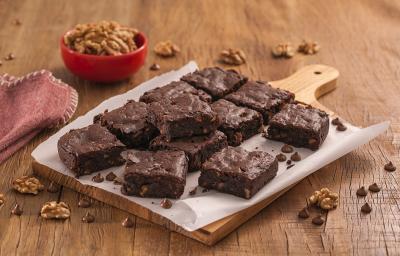 Fotografia em tons de marrom e vermelho de uma bancada de madeira com uma tábua e um papel manteiga, sobre ele pedaços de brownie. Ao fundo um recipiente vermelho pequeno com nozes e um pacote de brownie dois frades.