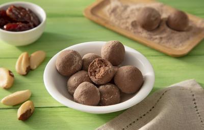 Fotografia em tons de marrom em uma bancada de madeira de cor verde. Ao centro, um bowl branco contendo as trufas. Ao lado, há algumas castanhas do pará e um pano marrom. Ao fundo, um pote com tâmaras e uma tábua com mais trufas.