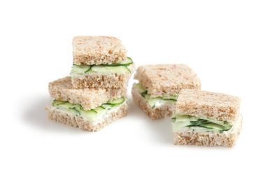 Fotografia em tons de verde em um fundo branco com quatro mini sanduichinhos no pão integral com recheio de pepino.