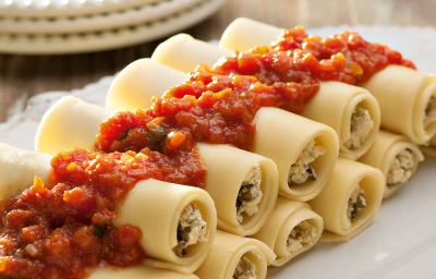 Fotografia em tons de vermelho em uma bancada de madeira com um recipiente oval branco grande com vários canelones de berinjela e com molho de tomate em cima dele.