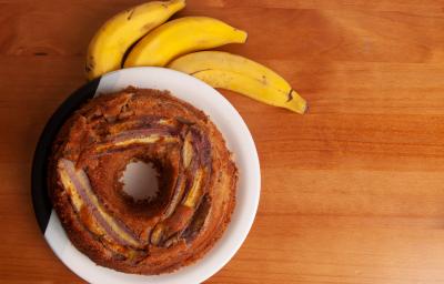 Fotografia em tons de marrom claro com um prato branco ao centro. Dentro do prato existe um bolo de banana redondo com um furo no meio, por cima do bolo existe pedaços de bananas caramelizadas. Ao lado existe três bananas inteiras.