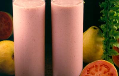 Fotografia em tons de rosa em uma bancada de madeira escura, dois copos de vidro alto com a bebida feita com suco de goiaba e Leite Moça. Ao lado, goiabas e uma goiaba aberta ao meio.