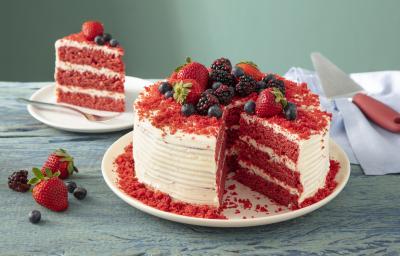 Fotografia em tons de vermelho em uma bancada de madeira clara, um prato redondo branco com o bolo red velvet cortado ao meio. Ao fundo, um prato pequeno com o pedaço.