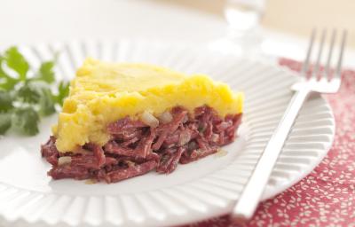 Fotografia em tons de vermelho em uma mesa de madeira clara com um prato redondo raso branco ao centro com uma fatia do escondidinho de carne seca com mandioquinha e coentro ao lado para enfeitar o prato.