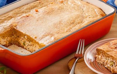 Fotografia em tons de creme, vermelho e azul, ao centro uma travessa vermelha onde a torta foi preparada e servida. Ao lado está uma fatia da torta em uma prato de sobremesa e um garfo.