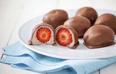 Fotografia em tons de branco de uma bancada branca, com um pano dobrado azul com bolinhas brancas, sobre ele um prato branco redondo com um bombom partido ao meio, mostrando o creme branco e o morango e cinco bombons inteiros.