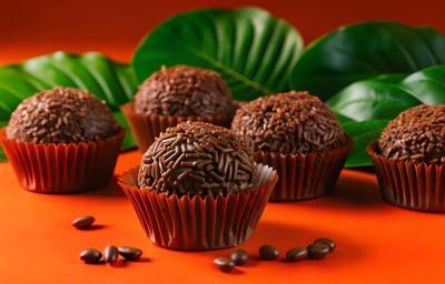 Fotografia de cinco brigadeiros passados no granulado, dentro de uma forminha vermelha, sobre uma mesa laranja com grão de café. No fundo tem algumas folhas.